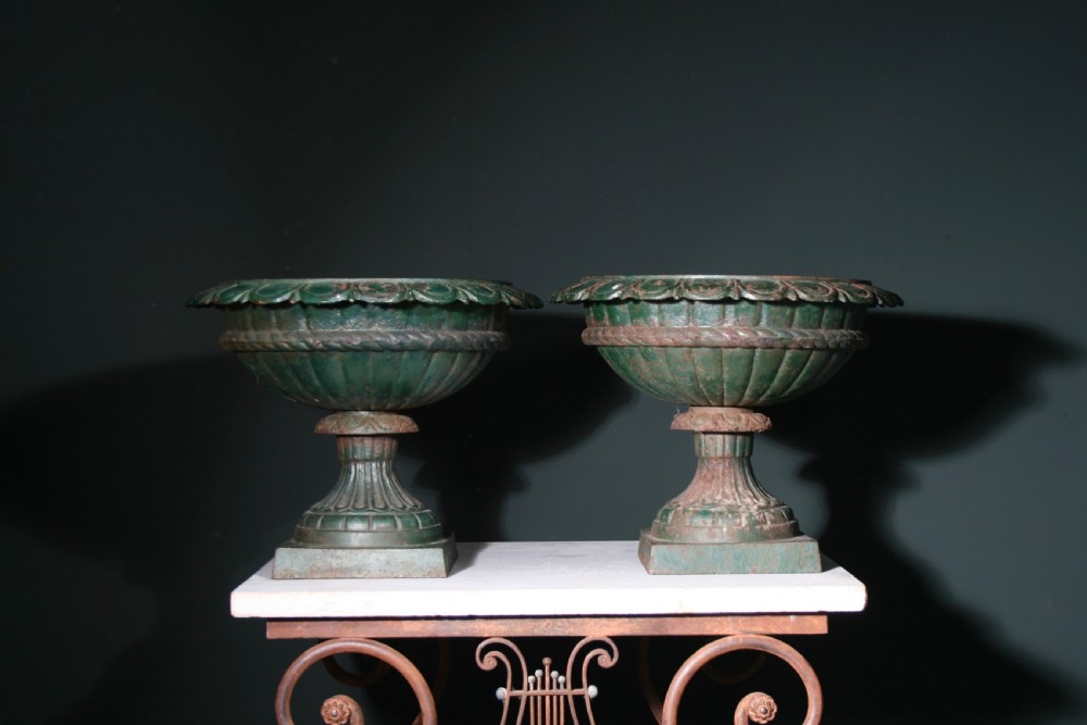 A pair of heavy cast iron garden urns featuring fluted bowls, scalloped rims, and square pedestal bases with weathered green paint and rust patina.