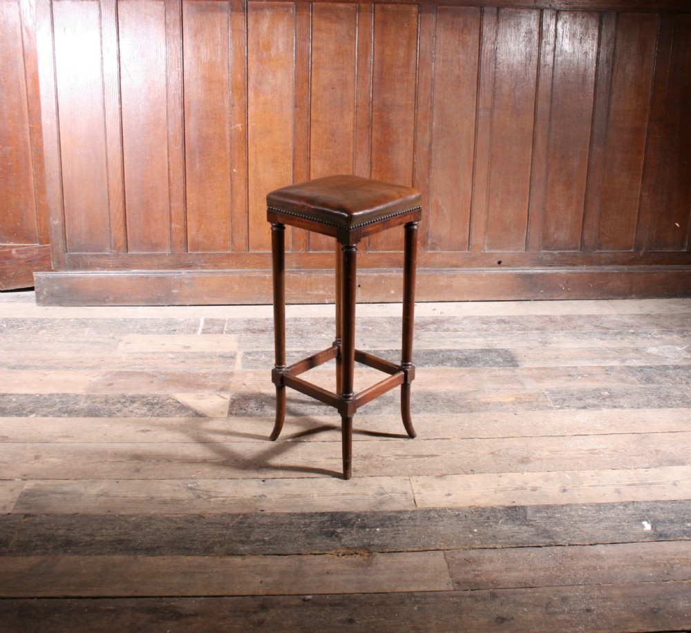 A tall antique wooden stool featuring a square padded brown leather seat with brass stud detailing and turned legs connected by stretchers.