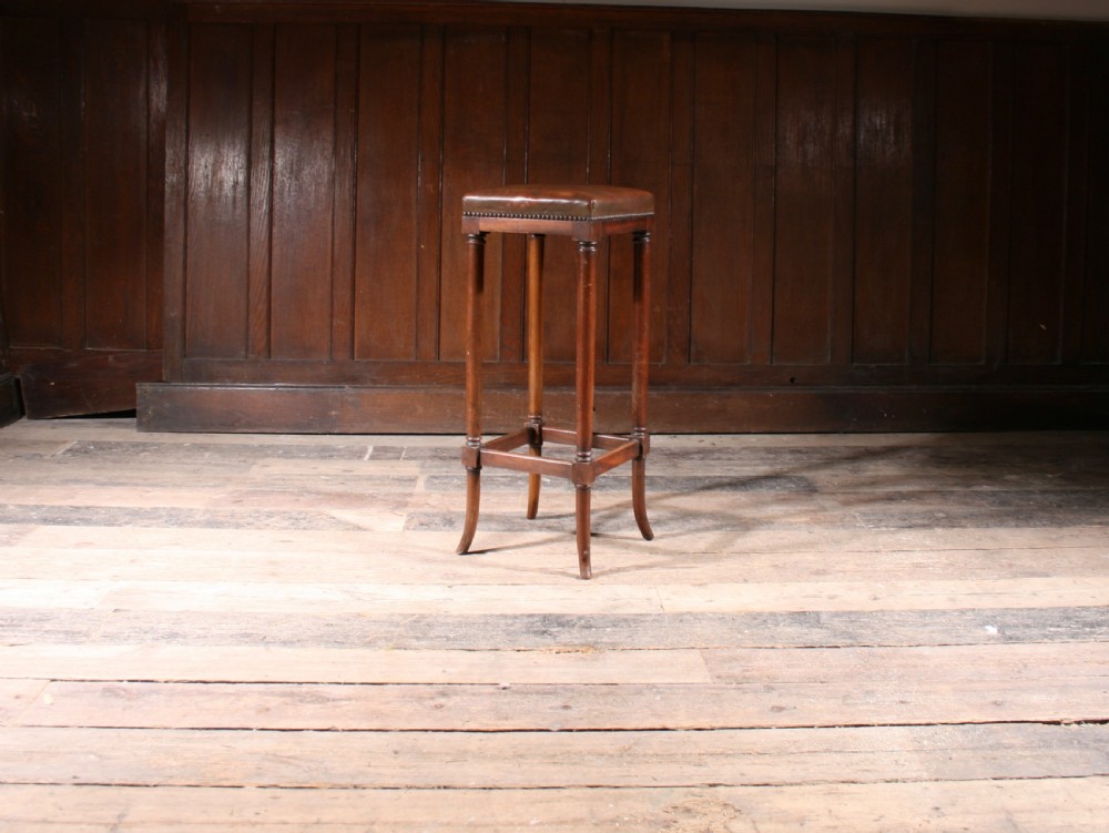 A tall antique wooden stool featuring a square padded brown leather seat with brass stud detailing and turned legs connected by stretchers. View II