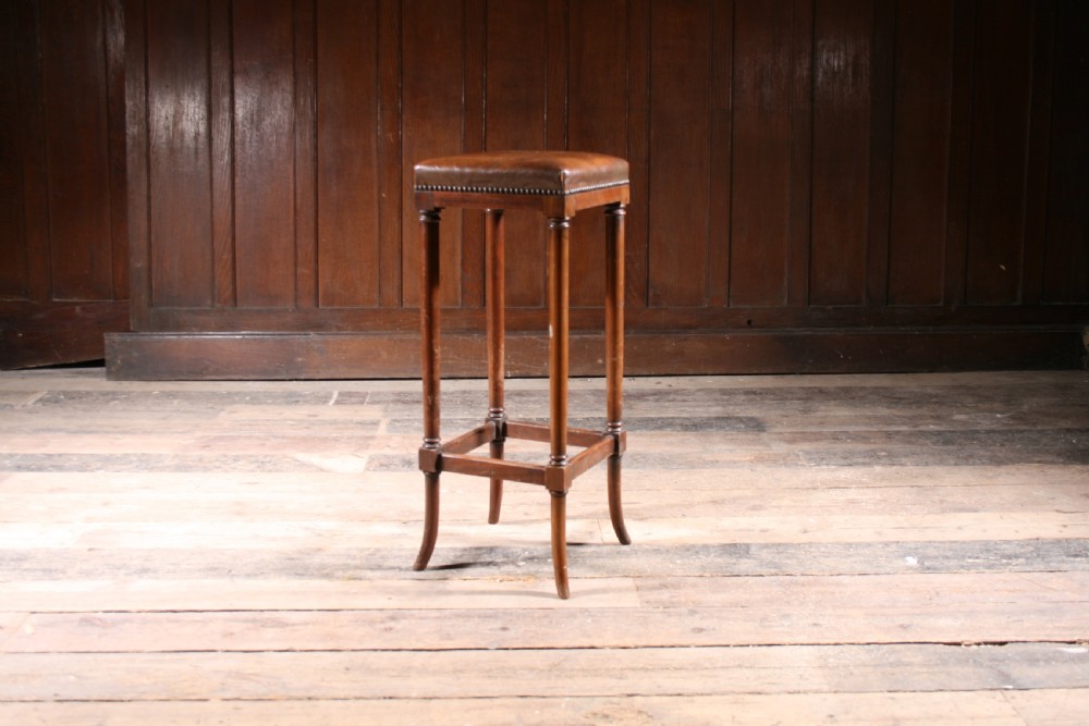A tall antique wooden stool featuring a square padded brown leather seat with brass stud detailing and turned legs connected by stretchers. View IV