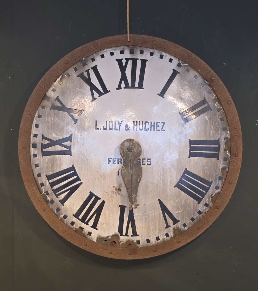 A large circular antique French turret clock face featuring black Roman numerals on a white enamelled surface with a riveted iron frame.