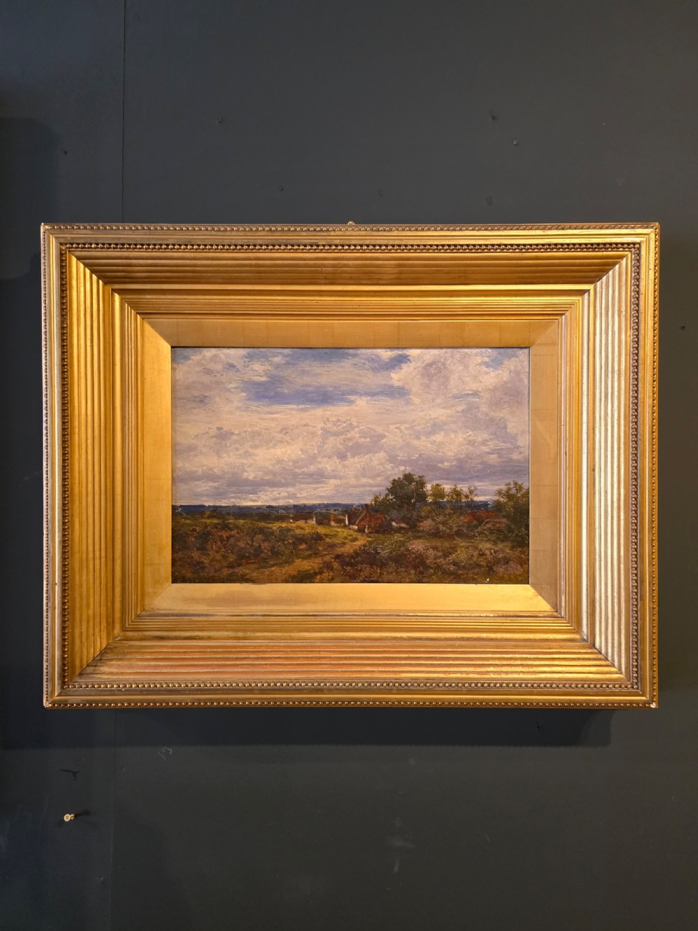 An 1896 Victorian landscape oil painting depicting an open rural field under a blue cloudy sky, encased in a gilded reeded wooden frame.