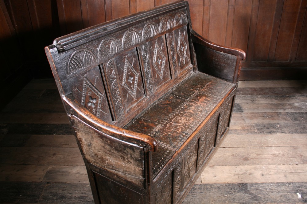A dark brown 17th-century carved oak settle with high panelled back featuring diamond lozenge carvings and a wooden bench seat. View IV