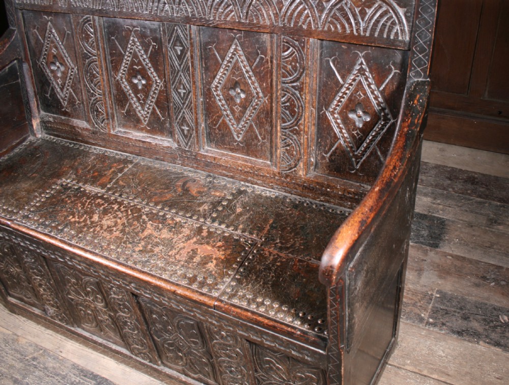 A dark brown 17th-century carved oak settle with high panelled back featuring diamond lozenge carvings and a wooden bench seat. View VII
