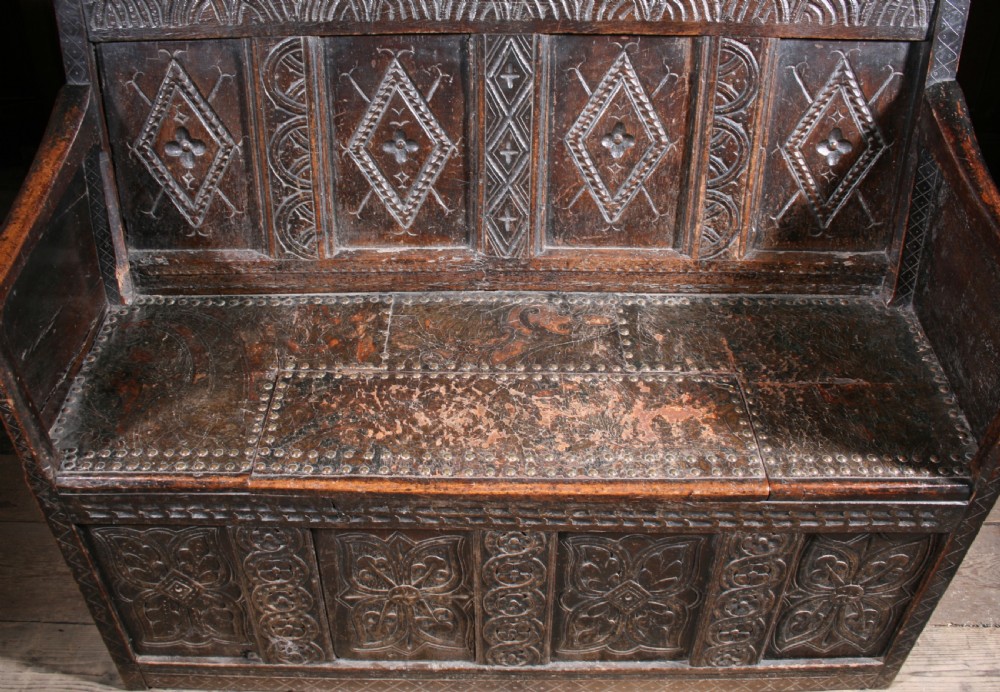 A dark brown 17th-century carved oak settle with high panelled back featuring diamond lozenge carvings and a wooden bench seat. View VIII