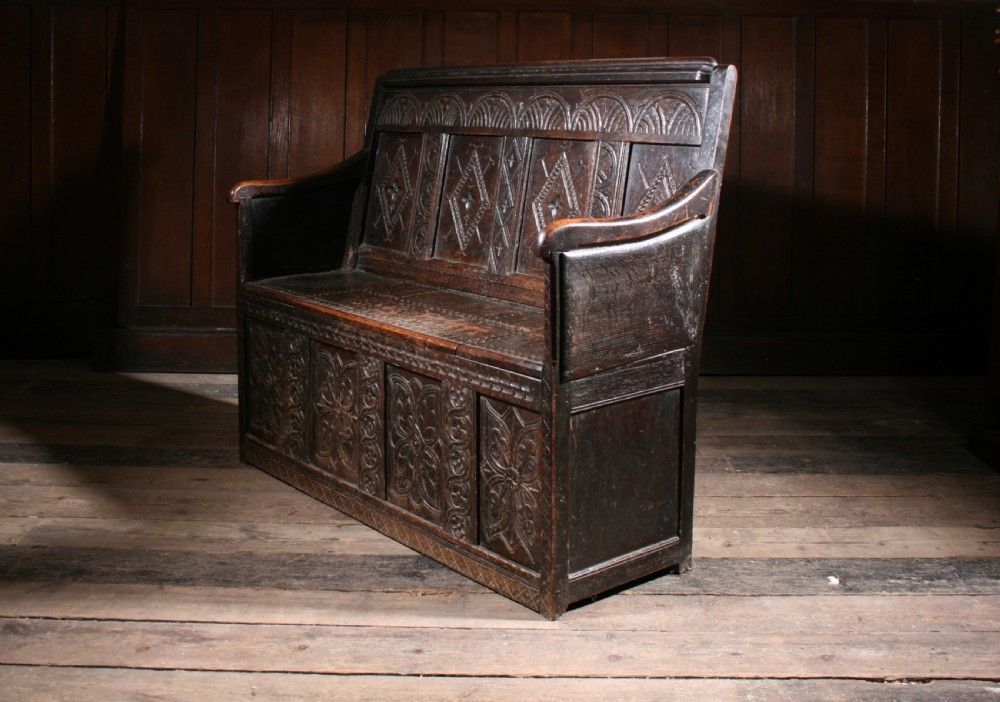 A dark brown 17th-century carved oak settle with high panelled back featuring diamond lozenge carvings and a wooden bench seat. View X