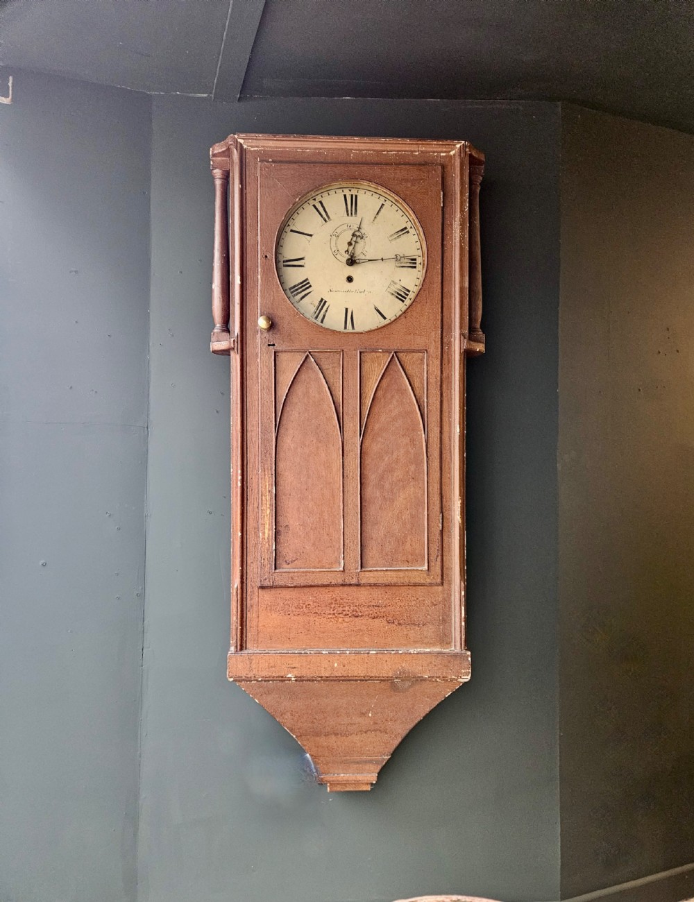 An antique pine wall clock featuring Gothic pointed arch panel details, turned side columns, and a circular white face displaying Roman numerals.