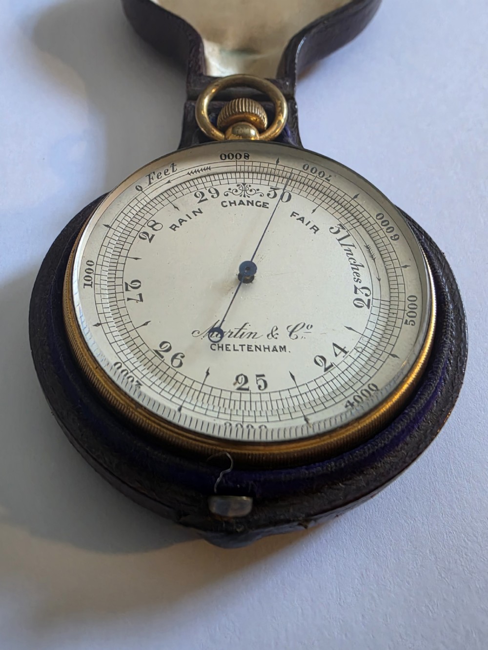 A circular Victorian brass pocket barometer with a silvered dial and altitude scale alongside its open plush-lined leather case. View II