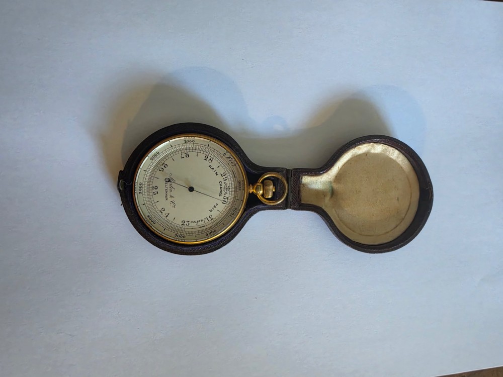 A circular Victorian brass pocket barometer with a silvered dial and altitude scale alongside its open plush-lined leather case. View IV