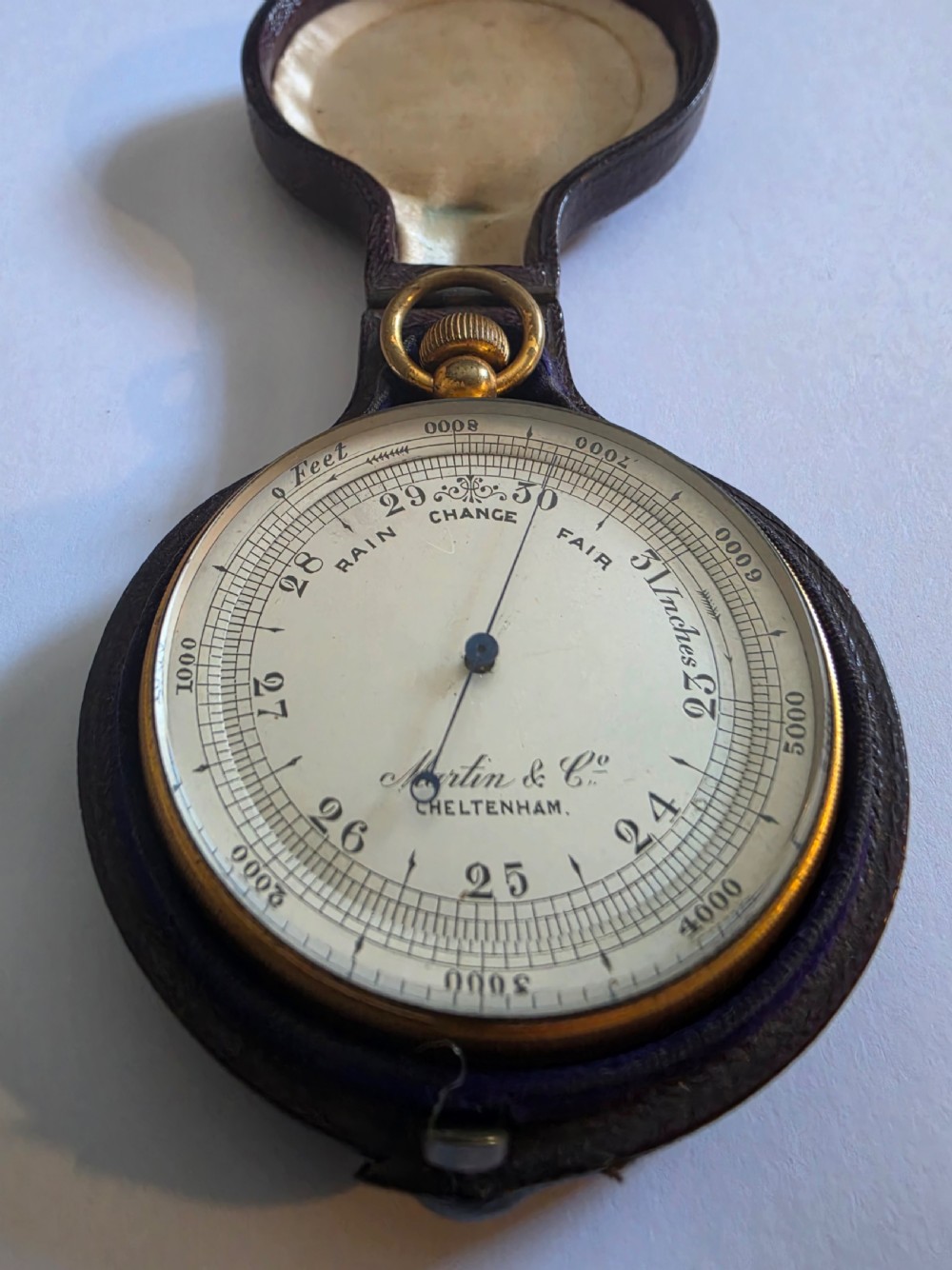 A circular Victorian brass pocket barometer with a silvered dial and altitude scale alongside its open plush-lined leather case. View VI