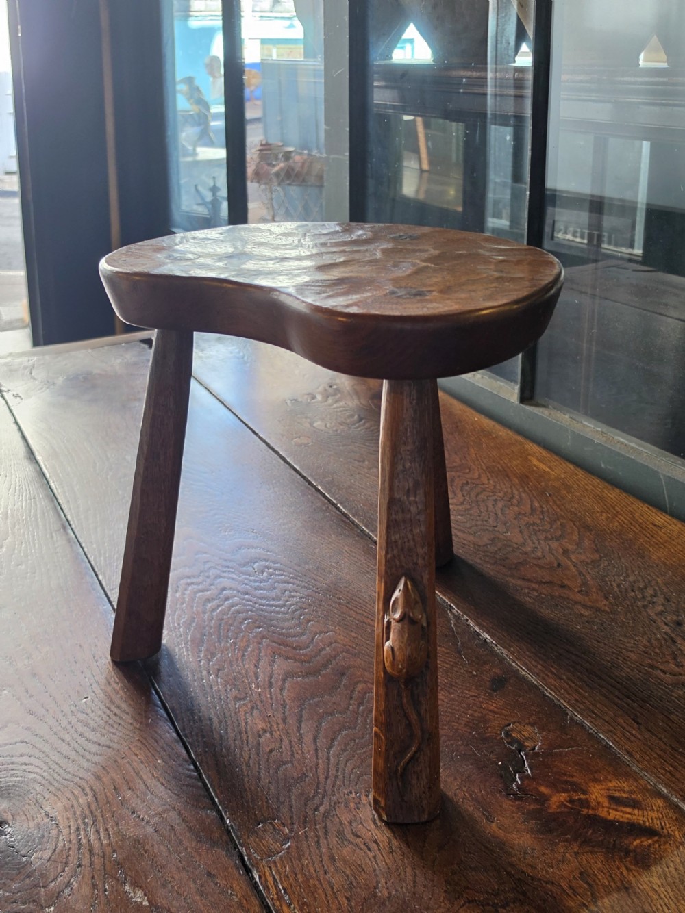 A dark English oak three-legged stool featuring an adzed kidney-shaped seat and a hand-carved mouse motif on one leg.