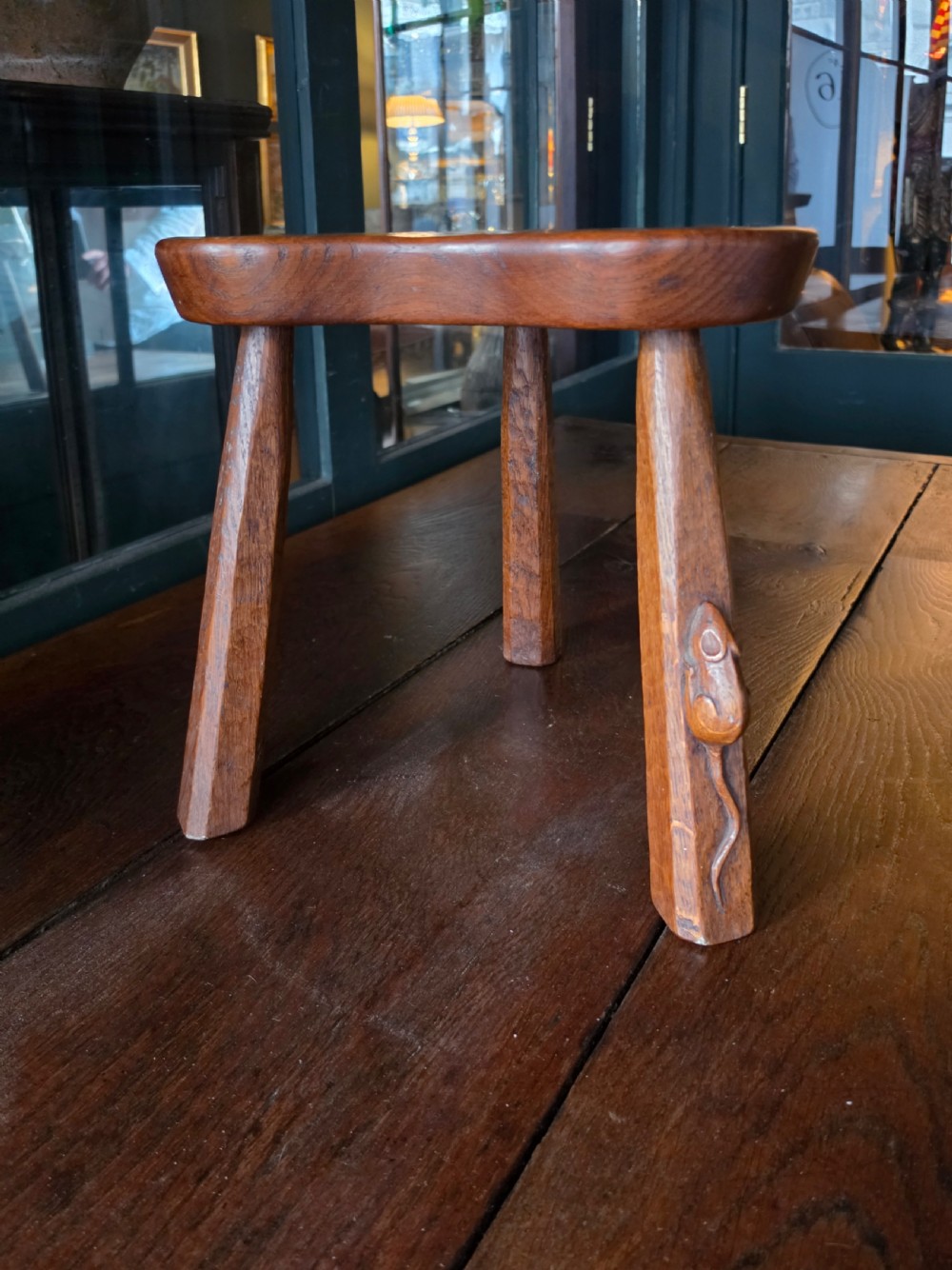 A dark English oak three-legged stool featuring an adzed kidney-shaped seat and a hand-carved mouse motif on one leg. View VIII