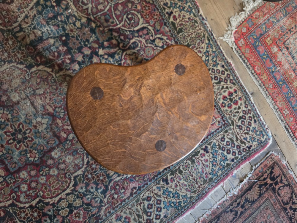 A dark English oak three-legged stool featuring an adzed kidney-shaped seat and a hand-carved mouse motif on one leg. View IX