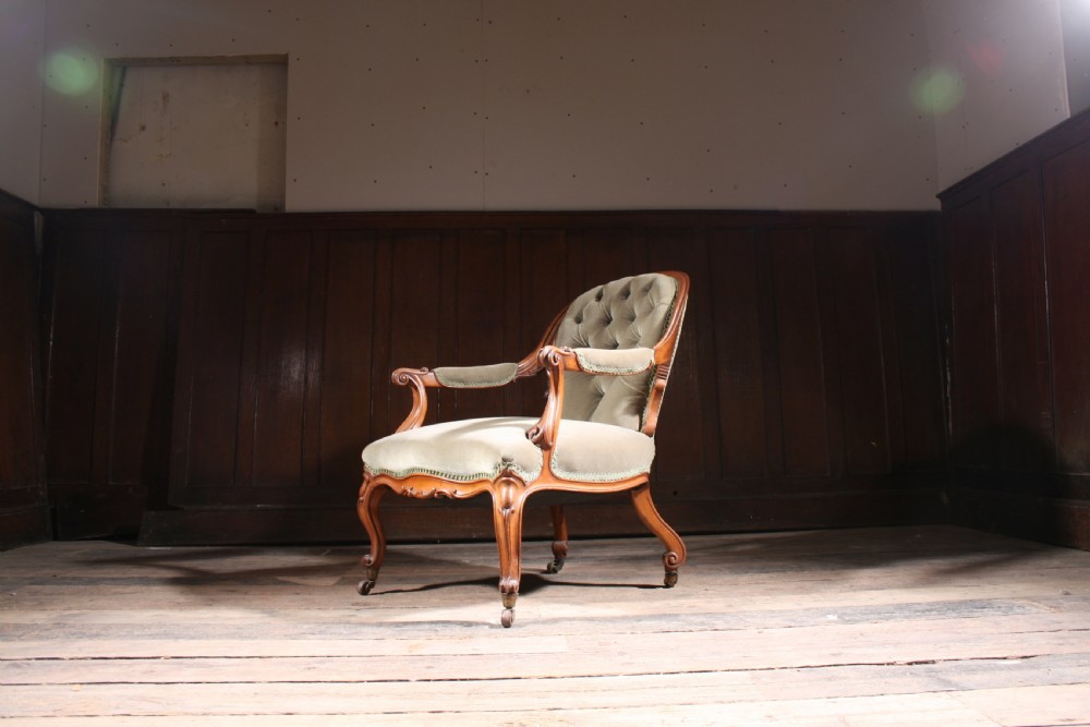 An antique walnut library armchair upholstered in button-tufted sage green velvet with carved cabriole legs on brass castors.