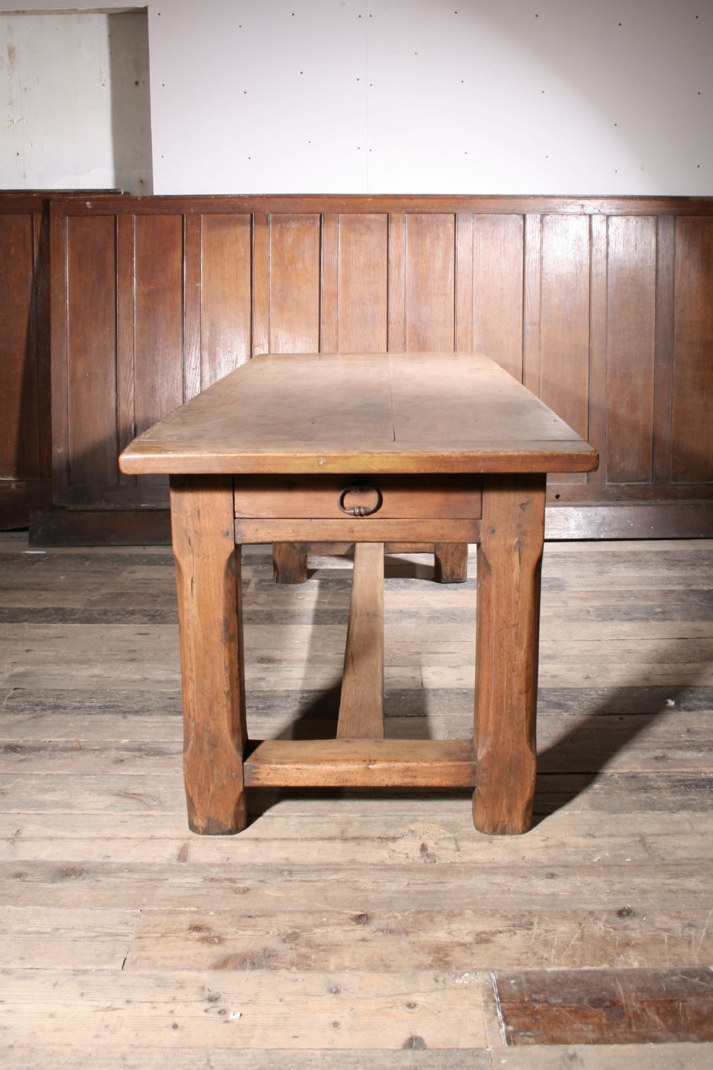 An antique walnut farmhouse dining table featuring a twin plank timber top, a single end drawer with a metal ring pull handle, and heavy legs connected by a central stretcher.