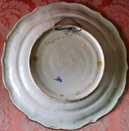 fig 1. A circular scalloped tin-glazed earthenware dish decorated with hand-painted figures in traditional attire set against a landscape, framed by a blue, green, and yellow scroll border with a heraldic crest at the top.