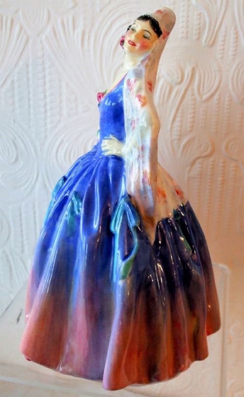 fig 1. A Royal Doulton porcelain figurine named Modena depicting a standing woman in a vibrant blue dress with a pink rimmed skirt and patterned head shawl.