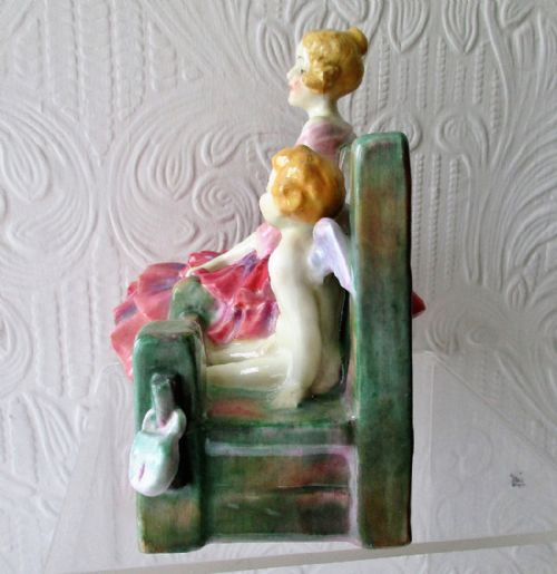 fig 1. A Royal Doulton porcelain figurine titled In the Stocks, featuring a seated lady in a ruffle-trimmed pink dress beside a winged cherub locked in wooden stocks.
