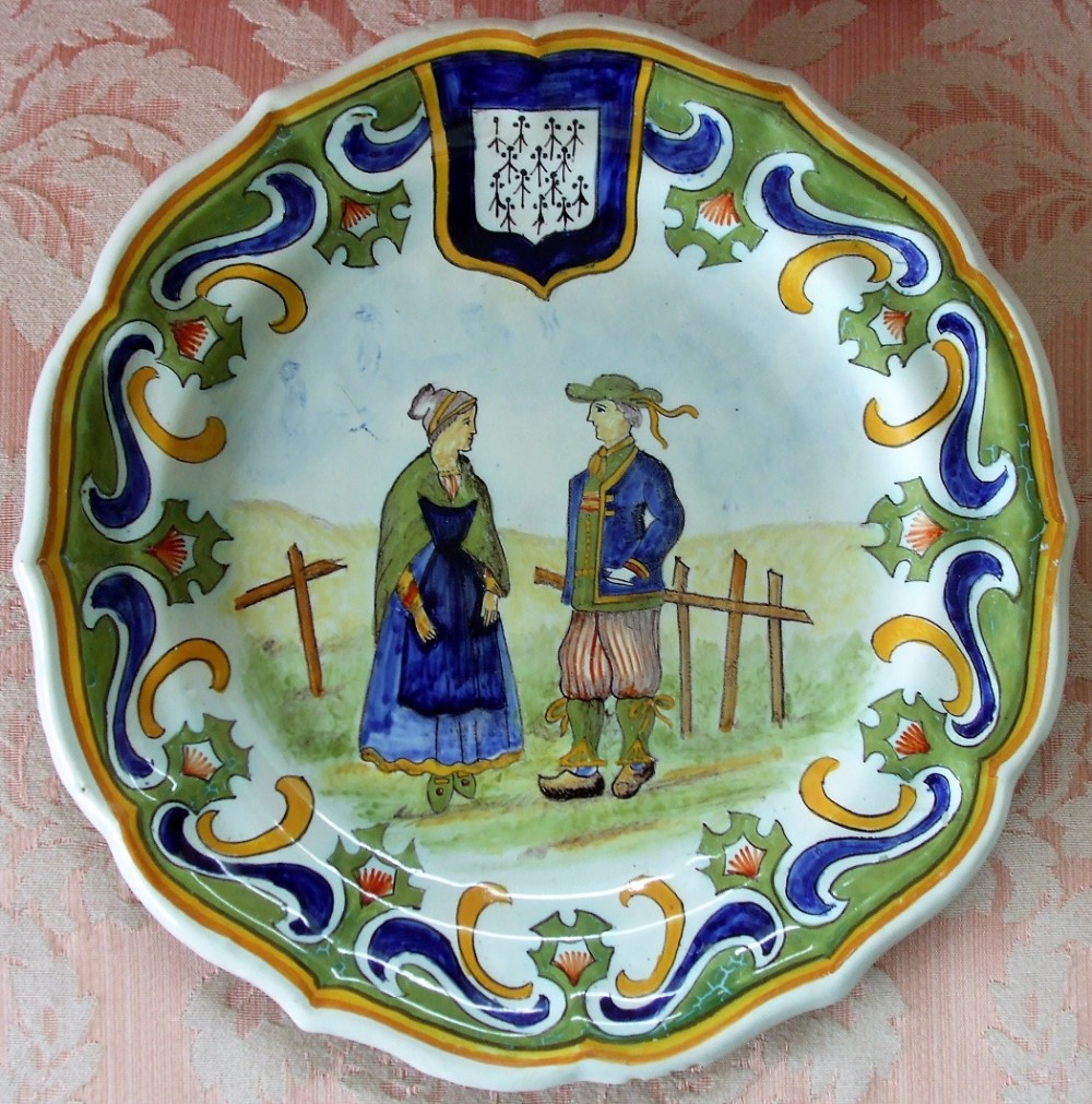 A circular scalloped tin-glazed earthenware dish decorated with hand-painted figures in traditional attire set against a landscape, framed by a blue, green, and yellow scroll border with a heraldic crest at the top.
