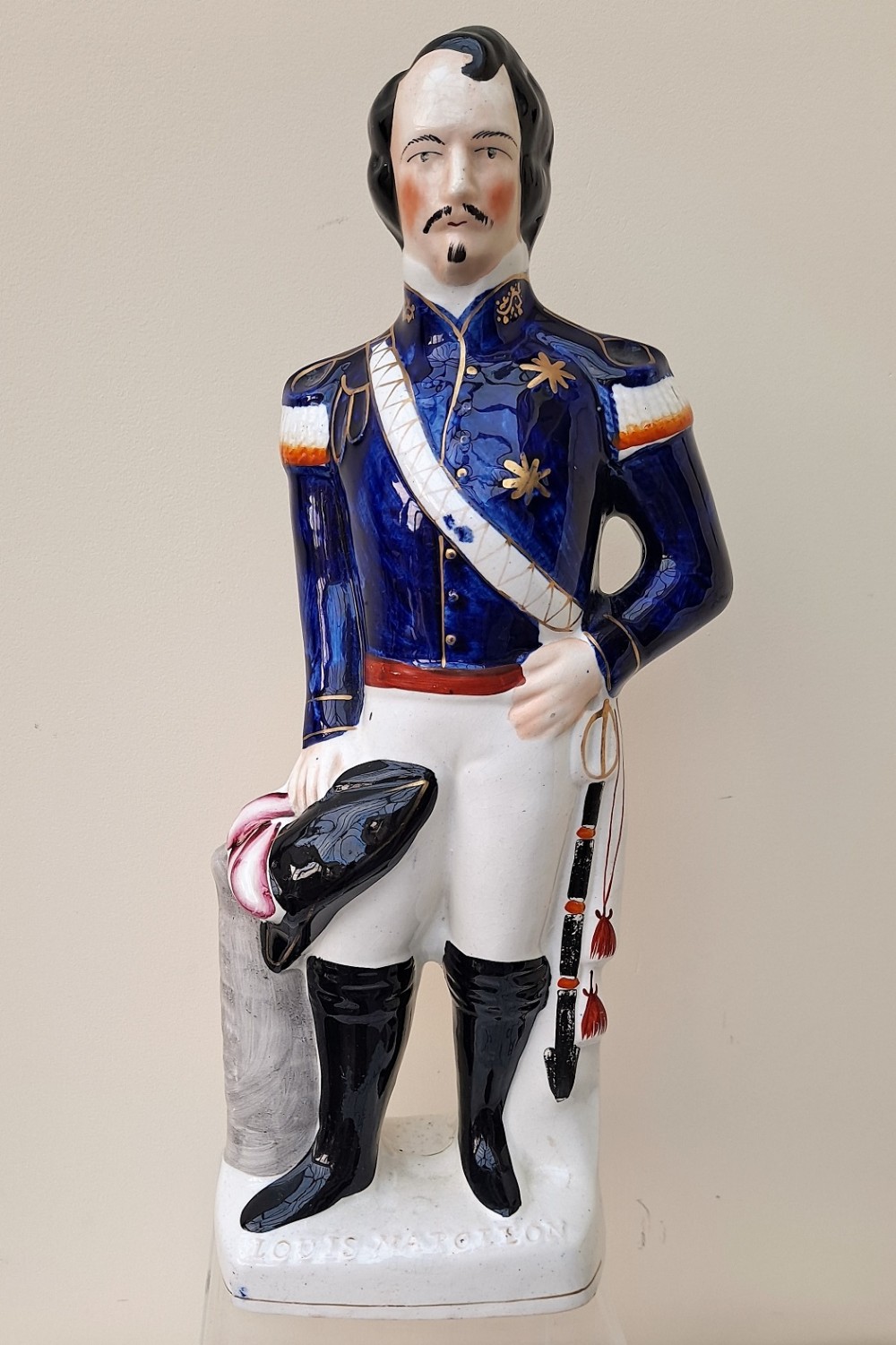 A Victorian Staffordshire earthenware figure depicting Louis Napoleon III in a cobalt blue military tunic, white trousers, black boots, and carrying a bicorne hat.