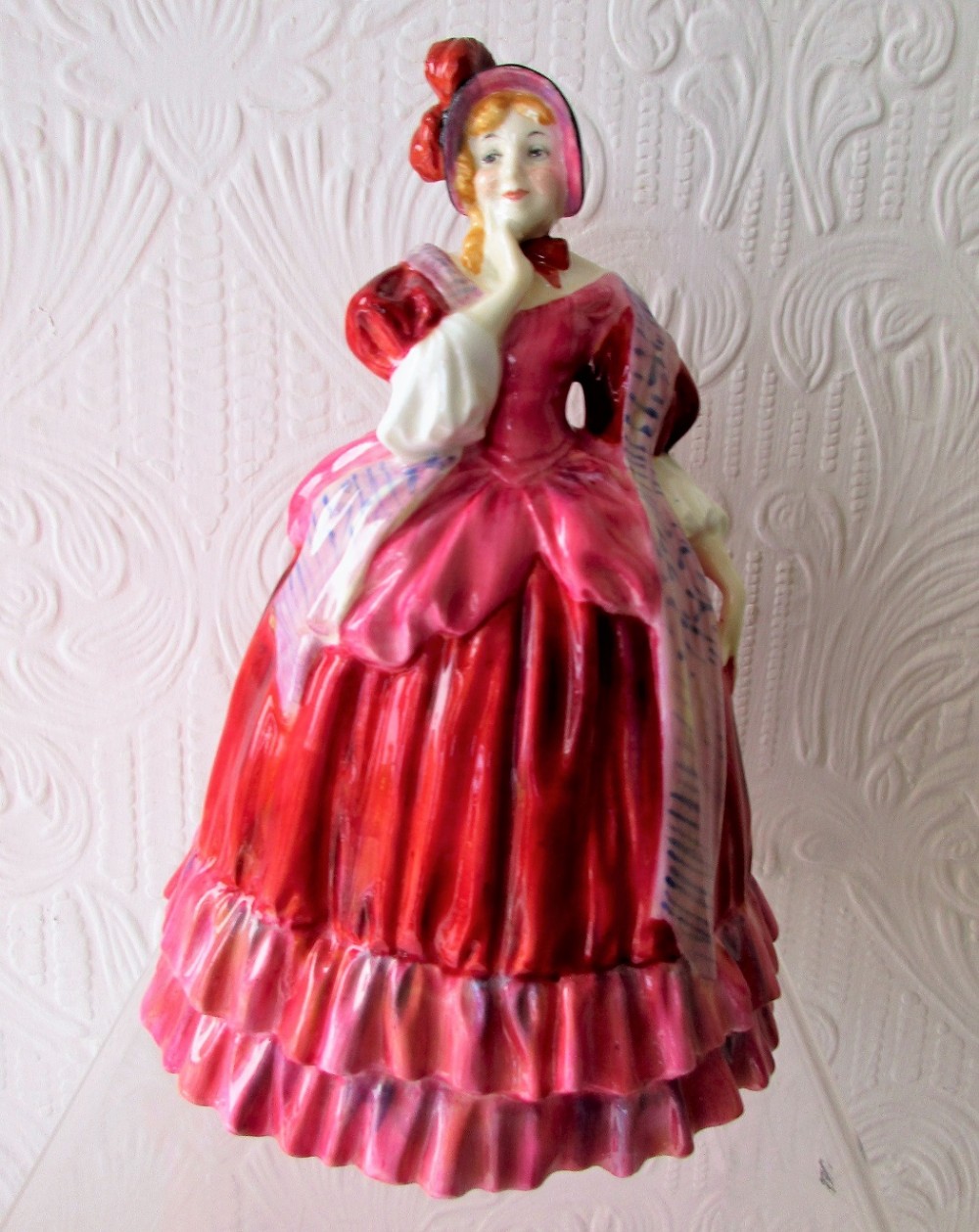 A vintage Royal Doulton glazed bone china figurine depicting a lady in a red bonnet and layered red and pink crinoline dress with a patterned shawl.