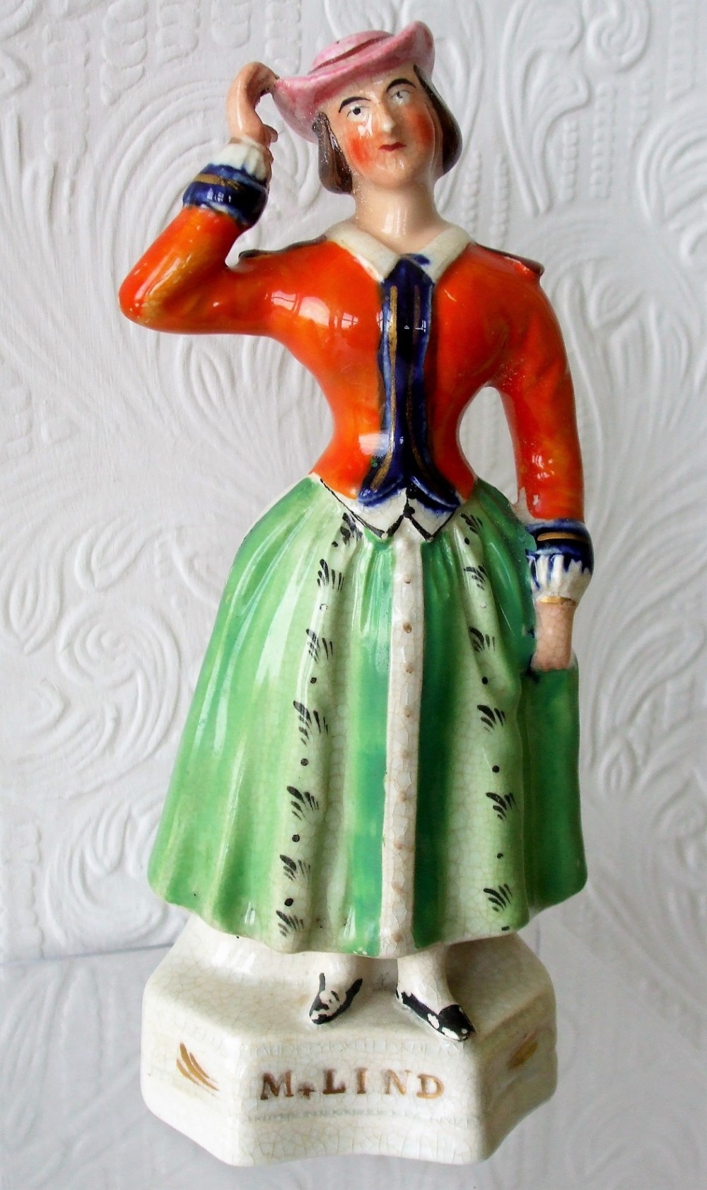 A Victorian Staffordshire earthenware figure of operatic performer Jenny Lind wearing an orange jacket, green skirt, and pink hat on a white base inscribed M. Lind.