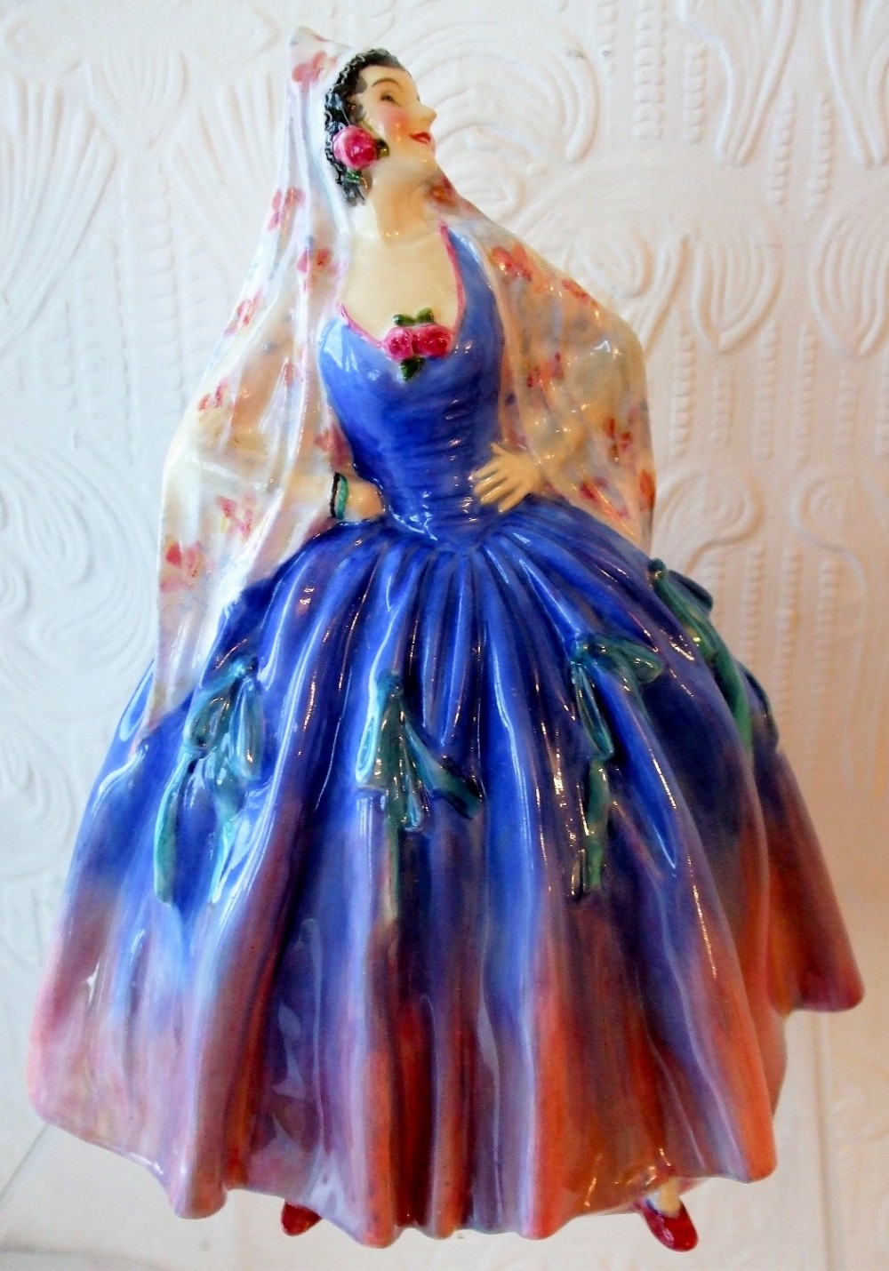 A Royal Doulton porcelain figurine named Modena depicting a standing woman in a vibrant blue dress with a pink rimmed skirt and patterned head shawl.
