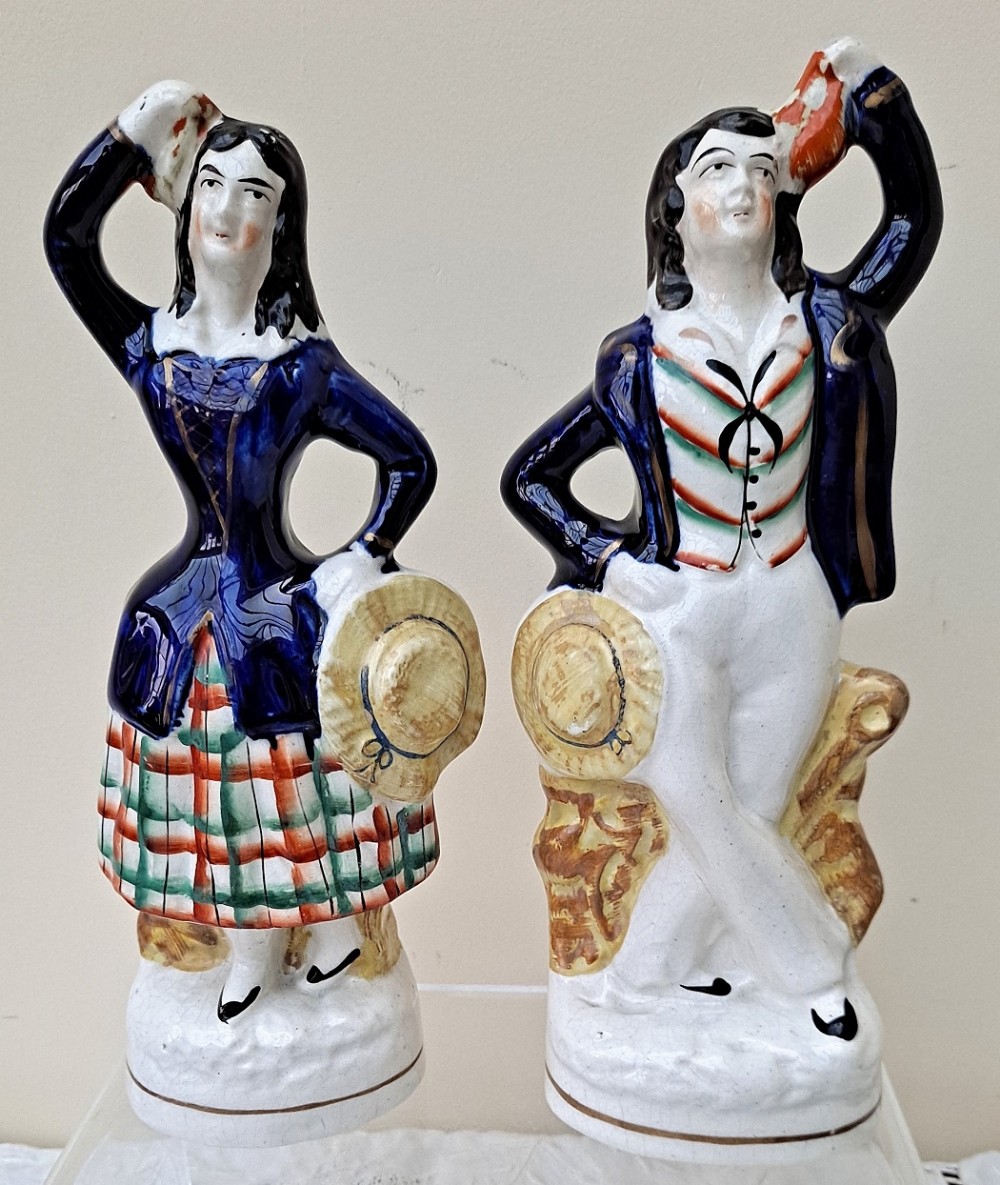 A pair of antique Victorian Staffordshire ceramic theatrical figures dressed in navy jackets, tartan patterned clothing, and holding hats.