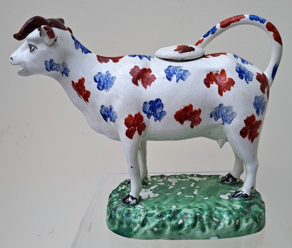 An antique white earthenware cow creamer with sponge-decorated blue and red patches standing on a moulded green base.