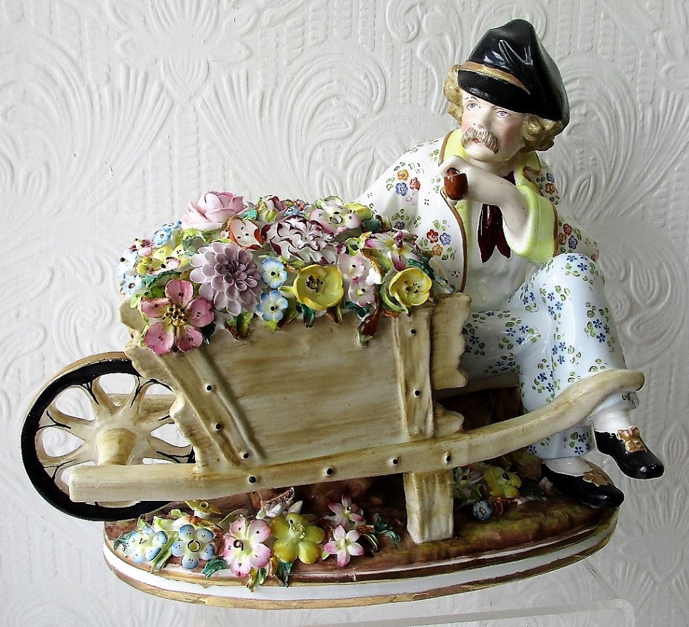 A Victorian hand-painted porcelain figurine depicting a seated flower seller with a wheelbarrow filled with hand-modelled blossoms.