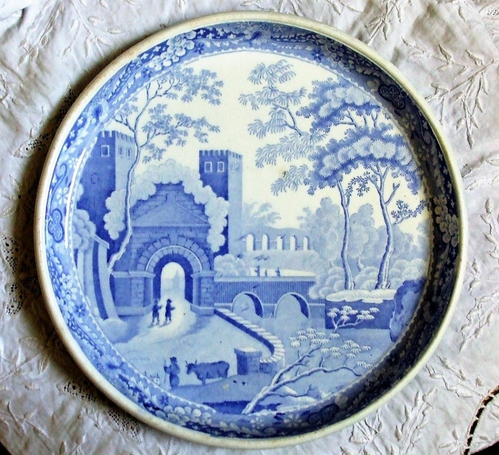 A circular Georgian blue and white ceramic tazza decorated with a printed scene of a castle archway, stone bridge, trees, and figures within a floral border.