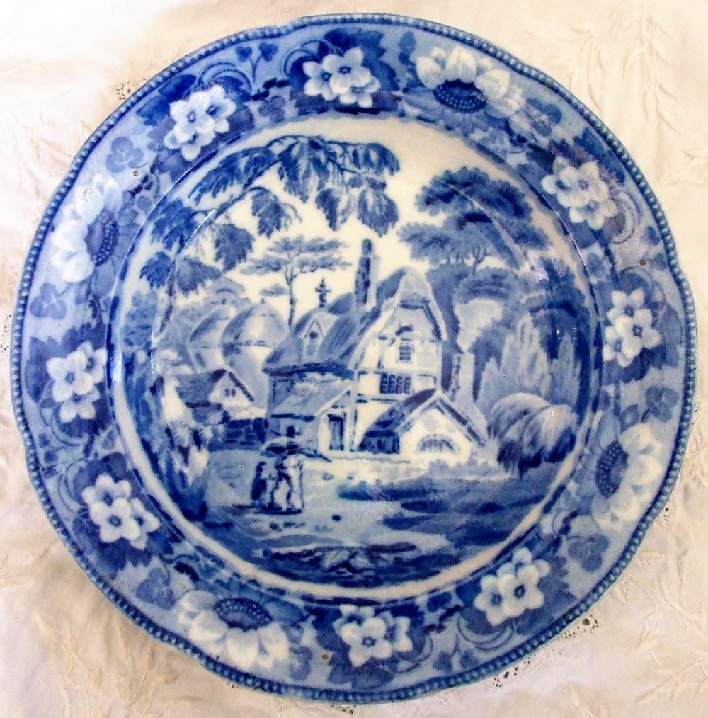 A circular Georgian blue and white earthenware plate featuring an underglaze transfer print of a rural cottage scene surrounded by a wide floral border.