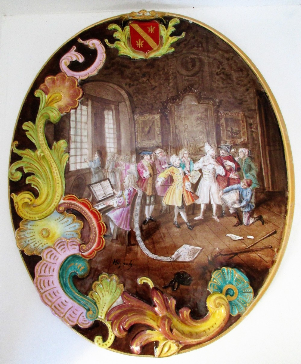 An oval French faience ceramic wall plaque featuring a hand-painted indoor genre scene surrounded by raised rococo acanthus scrolls and a coat of arms at the top.