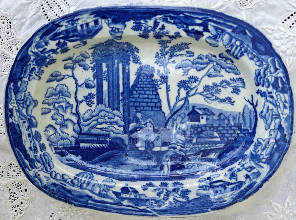 An oval Georgian earthenware serving dish featuring a cobalt blue transferware pattern of classical ruins and surrounding landscape.