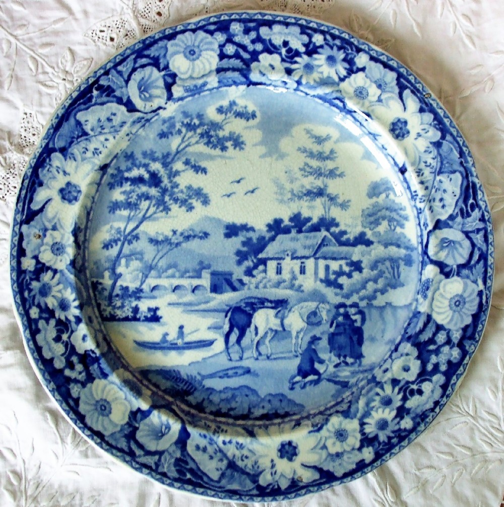 An antique Georgian circular earthenware plate featuring a cobalt blue and white transfer printed landscape with figures and horses near a cottage, surrounded by a floral border.