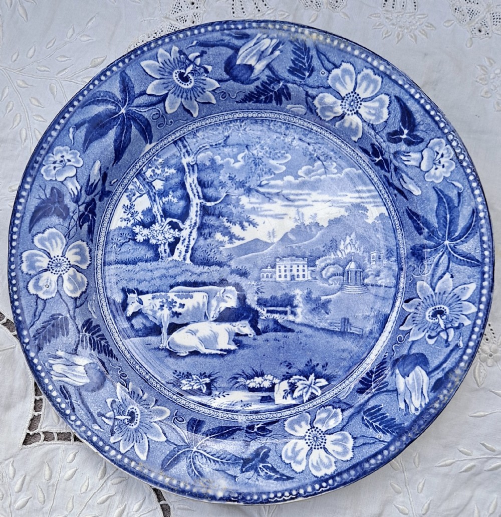 A circular Georgian blue and white transferware plate featuring a pastoral scene with cattle and a grand house surrounded by a detailed botanical border.