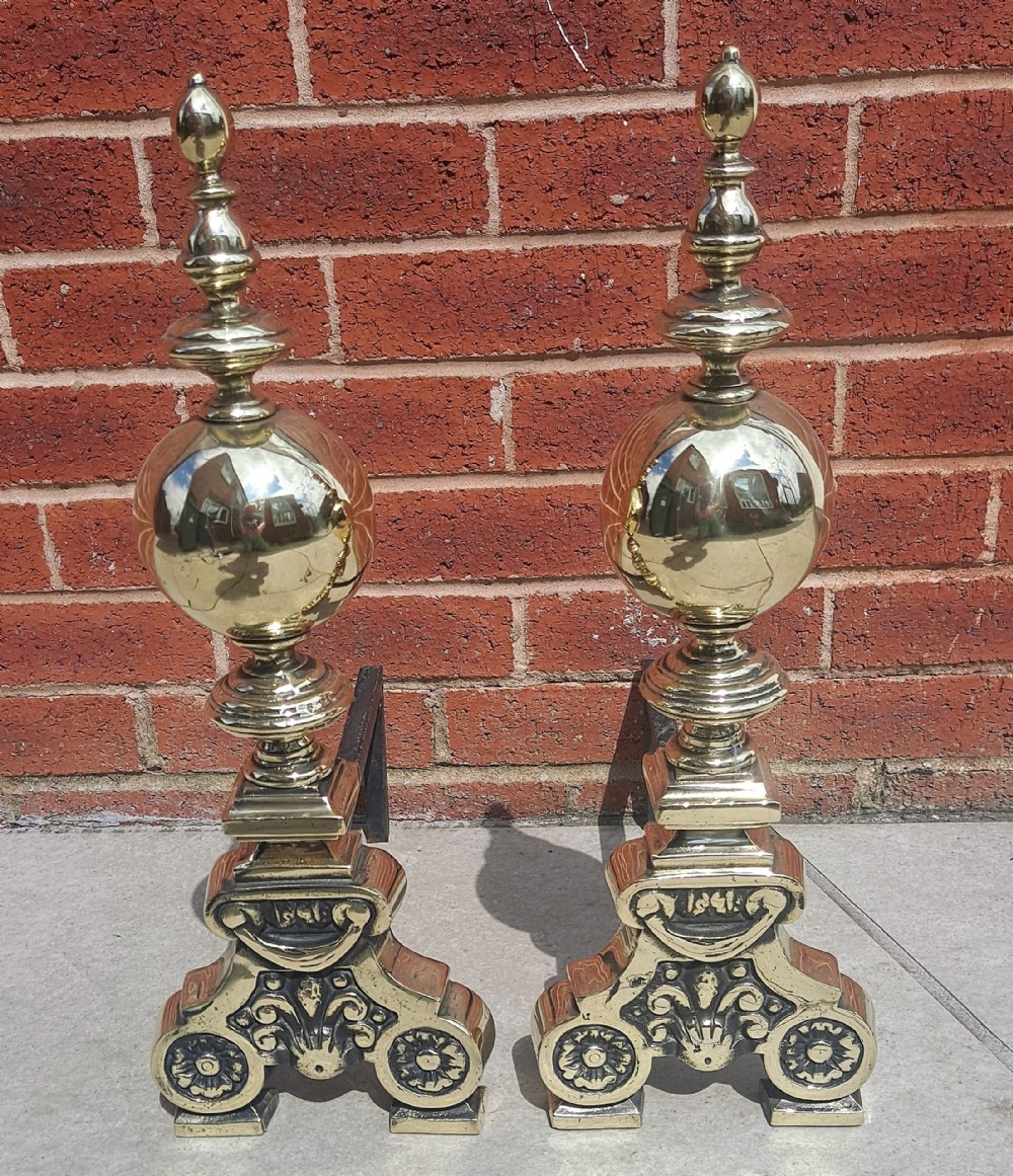A pair of antique Victorian polished brass andirons featuring large spherical finials mounted on ornate scrolled cast bases with iron rear billets.