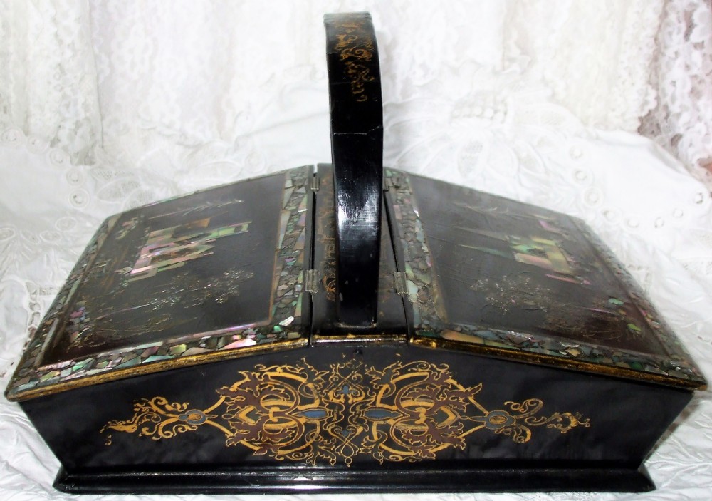 A Victorian black lacquered papier-mache needlework casket featuring mother-of-pearl inlay, gilded foliate patterns, and an overhead carrying handle.