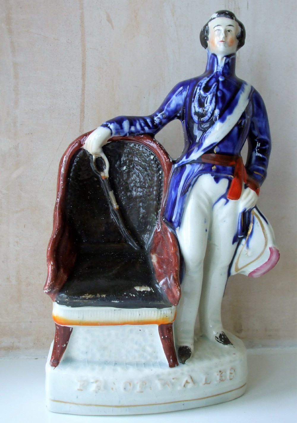 An antique Victorian Staffordshire earthenware portrait figure depicting the Prince of Wales in a cobalt blue jacket standing beside a chair on an inscribed white base.