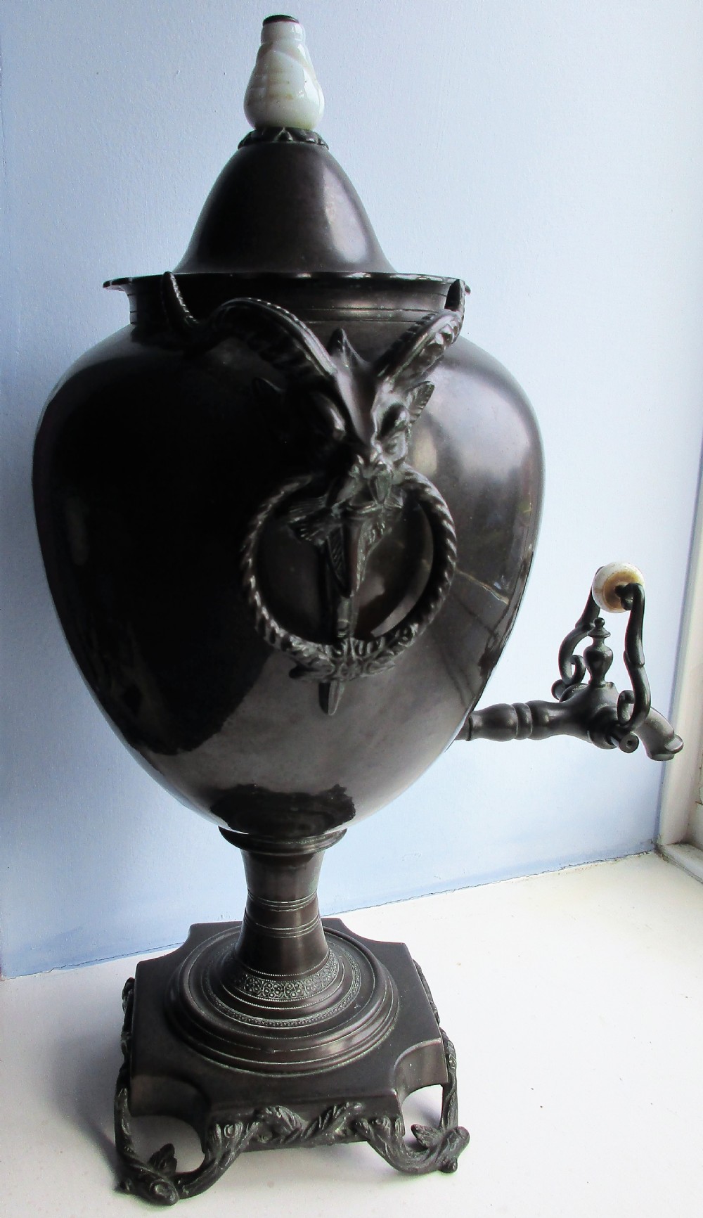 An antique Victorian japanned dark metal tea urn featuring a tapered body, ornate cast side handles, a front spigot, and a white top finial. View IV