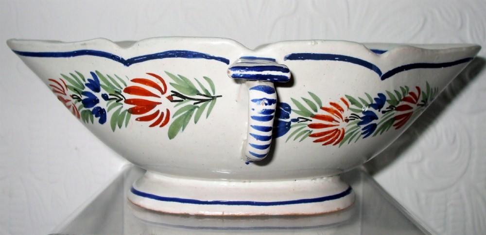 An off-white tin-glazed earthenware double sauceboat featuring hand-painted red and blue botanical decorations and a scalloped blue rim.