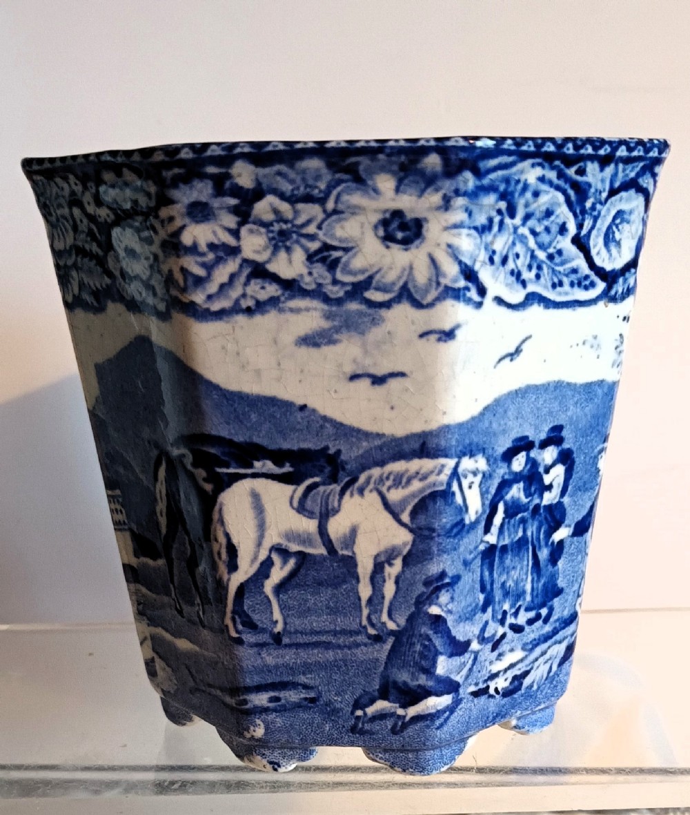 An octagonal blue and white ceramic transferware cachepot featuring a rural landscape scene with horses and figures framed by a floral border.