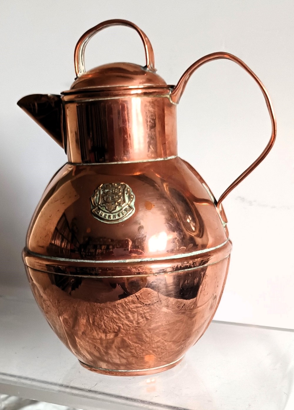 A bulbous Victorian polished copper Guernsey milk can featuring a fitted lid, loop handles, and a raised brass Guernsey crest medallion.