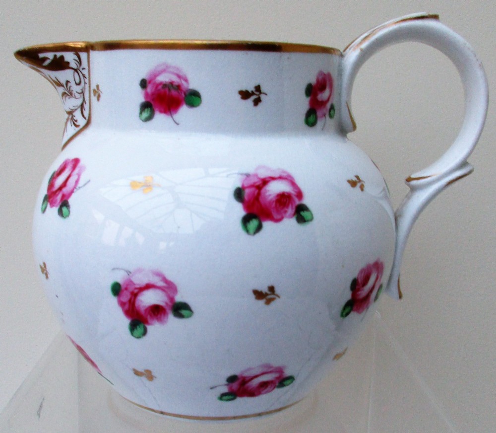 An antique white porcelain cream jug featuring hand-painted pink rose sprigs, gold leaf detailing, a pointed sparrow beak spout, and a curved handle.