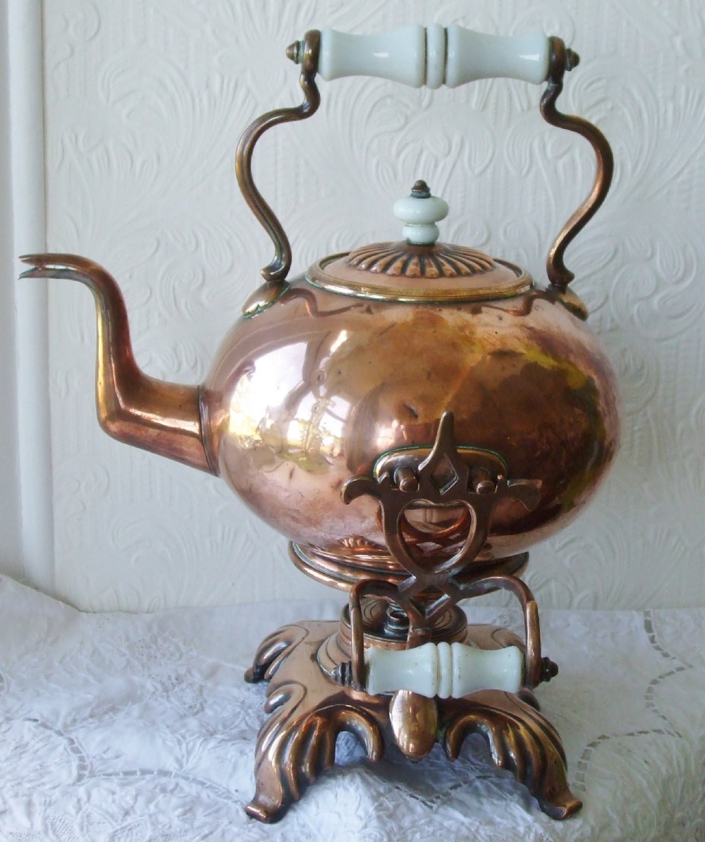 An antique Victorian copper spirit kettle featuring a white ceramic handle, gooseneck spout, and decorative brass stand with burner.
