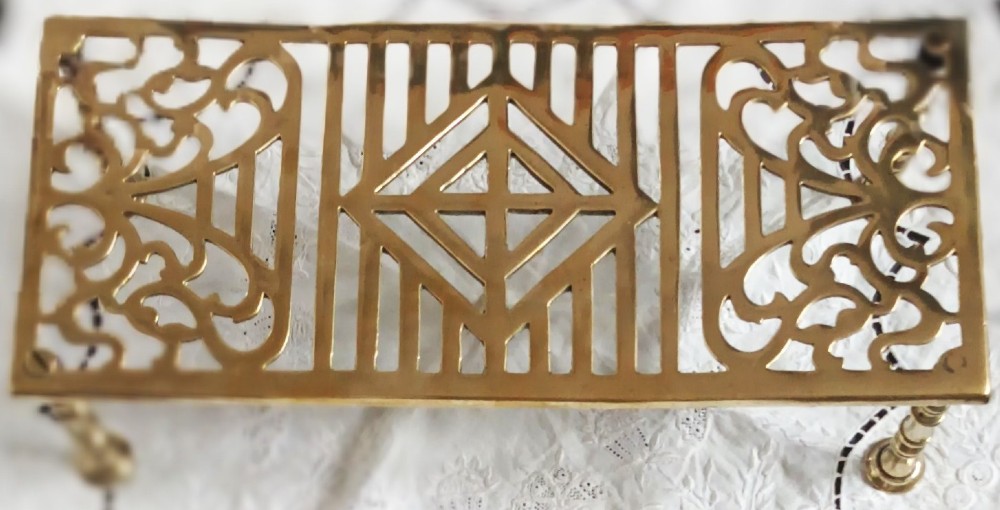 A rectangular polished brass trivet featuring openwork geometric and scroll fretwork supported on four turned legs. View III