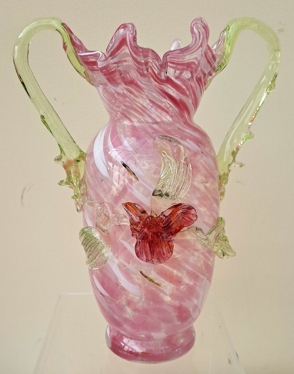 A Victorian hand-blown glass vase featuring a pink swirled body, a ruffled rim, an applied red flower accent, and two green vaseline glass handles.