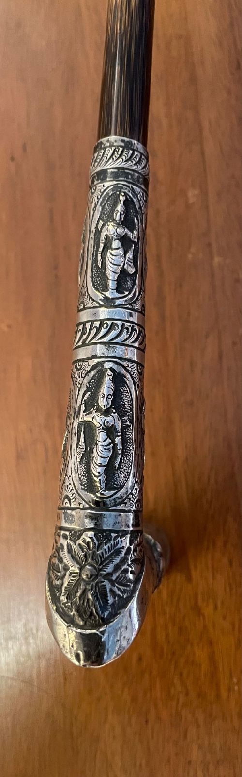 An antique walking stick with an embossed L-shaped silver handle featuring figurative motifs mounted on a dark brown bamboo shaft. View V