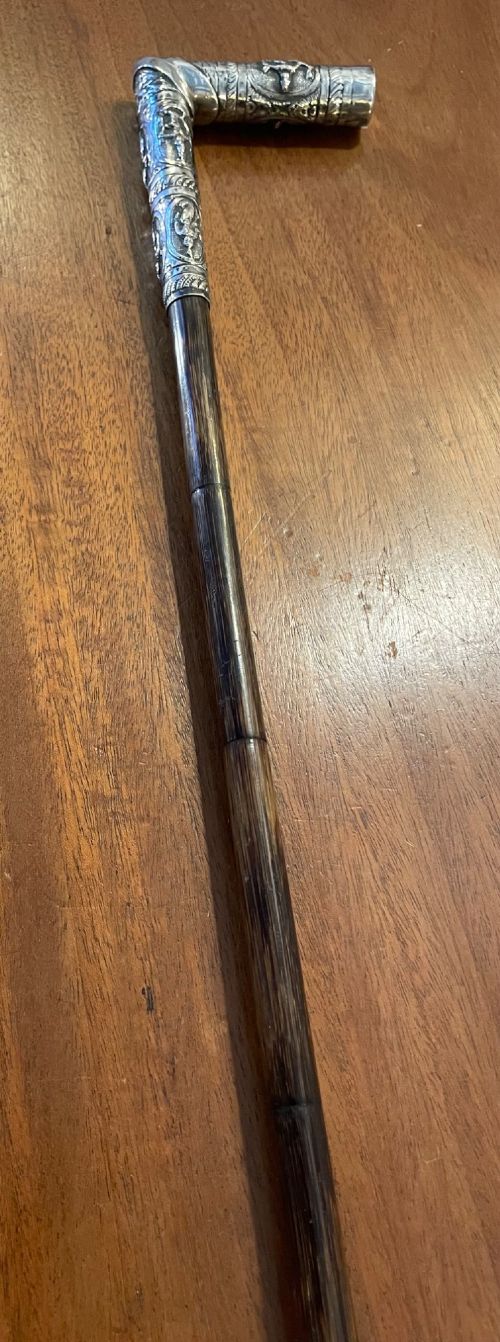 An antique walking stick with an embossed L-shaped silver handle featuring figurative motifs mounted on a dark brown bamboo shaft. View VII