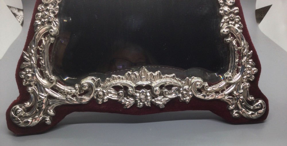 A late Victorian sterling silver easel mirror with embossed floral scrollwork, bevelled glass, and burgundy velvet backing. View VIII